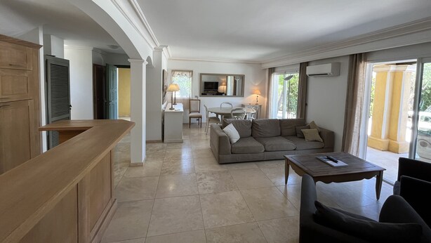 A spacious living area with a sofa, a coffee table, and a dining table. There is a kitchen in the background with a TV and a lamp.