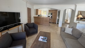 A living room with a flat-screen TV, a sofa, a coffee table, and a kitchen area with wooden cabinets and a central island.