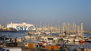 A marina with numerous boats docked, a large ship in the background, and a lighthouse to the right.