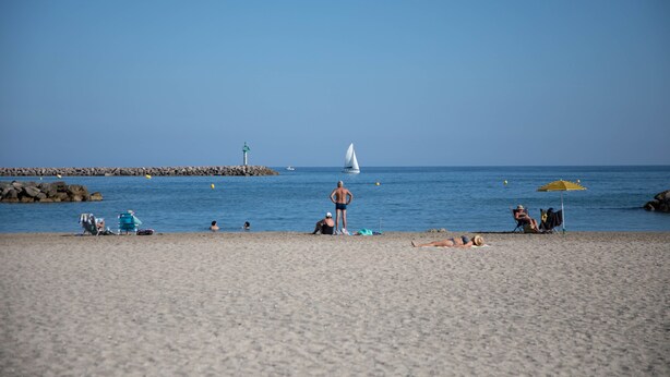 A beach with people relaxing, a sailboat, and a lighthouse in the distance.