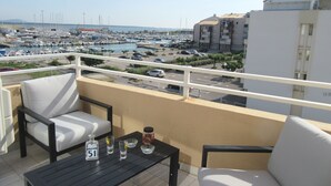A balcony with white cushioned chairs and a table, overlooking a marina with boats and a building in the background.