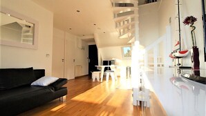 A modern living room with a black sofa, a white dining table, and a staircase leading to an upper level. - COUP DE ❤️ LA MOUETTE DEAUVILLAISE🐦classified 4☆ Tennis-Pool-Parking-Linen-Wifi (DEAUVILLE)