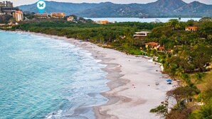 A beach with clear turquoise waters, a sandy shore, and a backdrop of mountains and buildings. - 5mins to Marina-Oceanview-Full Kitchen-Pool-AC-Gym – Presidential Suites 4 (Playa Flamingo)