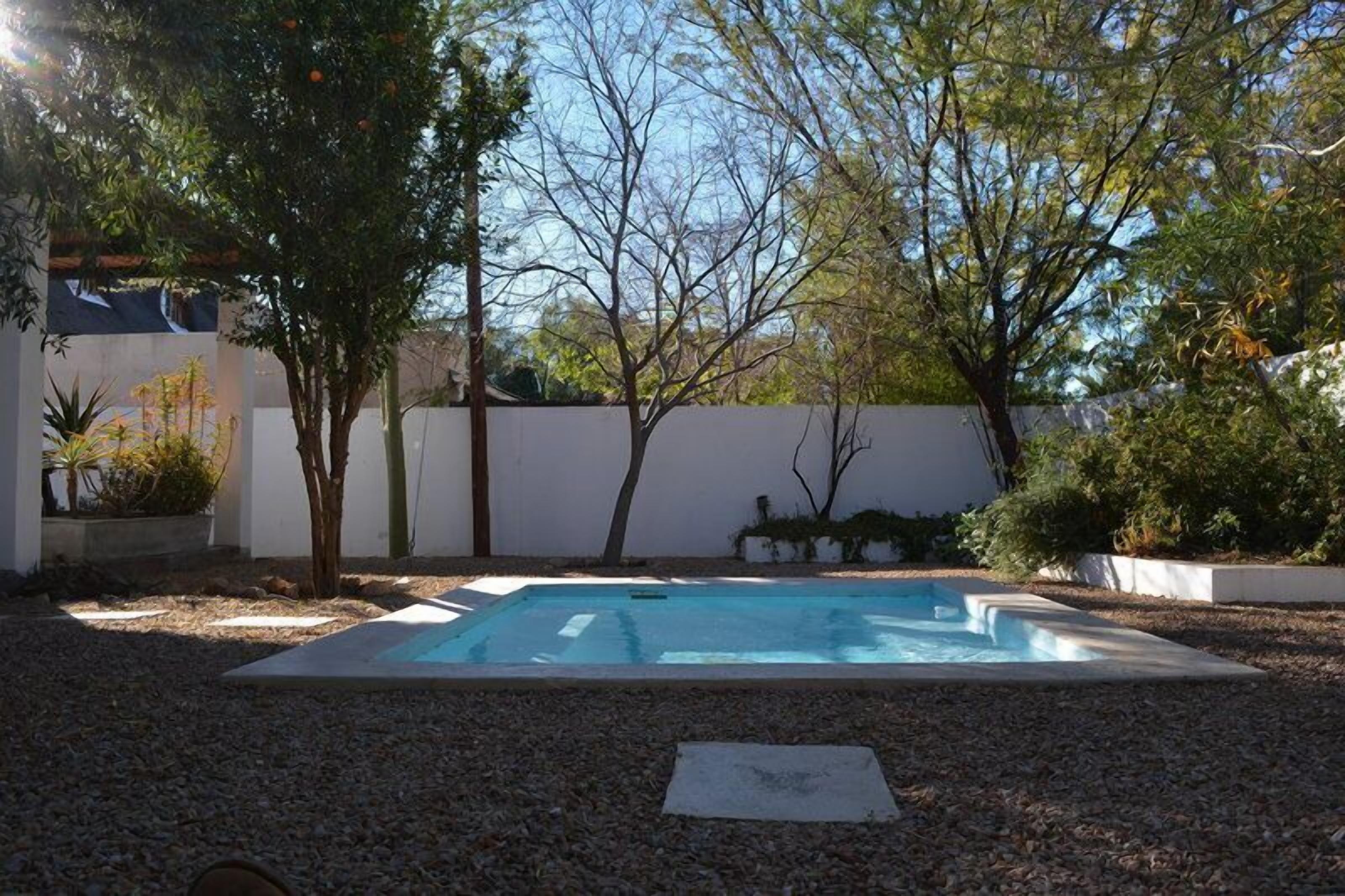 A rectangular swimming pool surrounded by gravel, with trees and a white wall in the background.