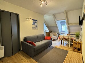Un salon chaleureux avec un canapé gris, une petite table et un fauteuil en osier. Une affiche est accrochée au mur, et une porte est visible sur la gauche.
