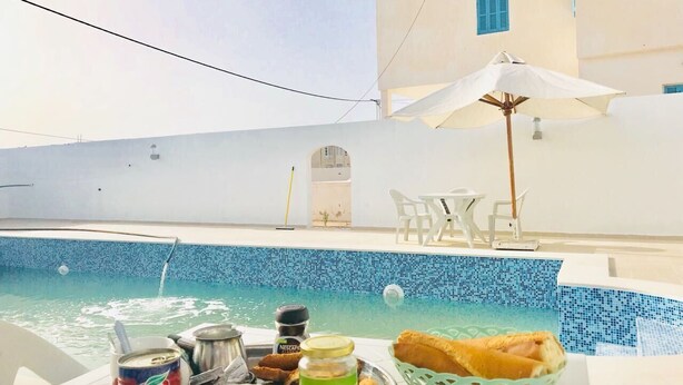 A poolside dining area with a table set for two, a white umbrella, and a view of a building with blue shutters. - Dar AESHA (Without Vis à Vis) (Djerba)