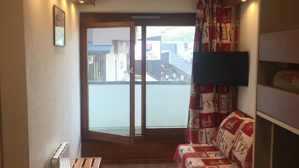 Ein kleiner Raum mit einer hölzernen Bank, einem Sofa mit gemusterten Kissen, einem Fernseher und Blick auf ein Gebäude durch ein großes Fenster.