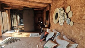 Une chambre rustique avec un lit, un mur décoré d’objets tissés et une vue sur l’extérieur.