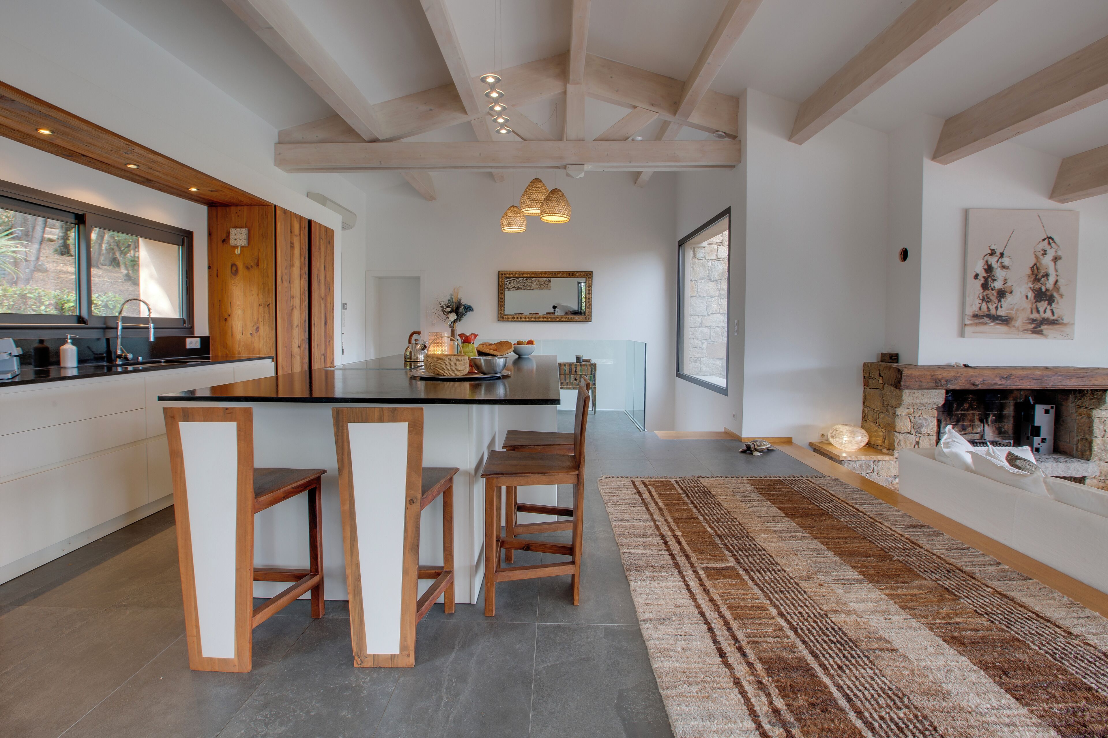 A modern kitchen with wooden accents, a central island with bar stools, a stone fireplace, and a large window providing natural light.