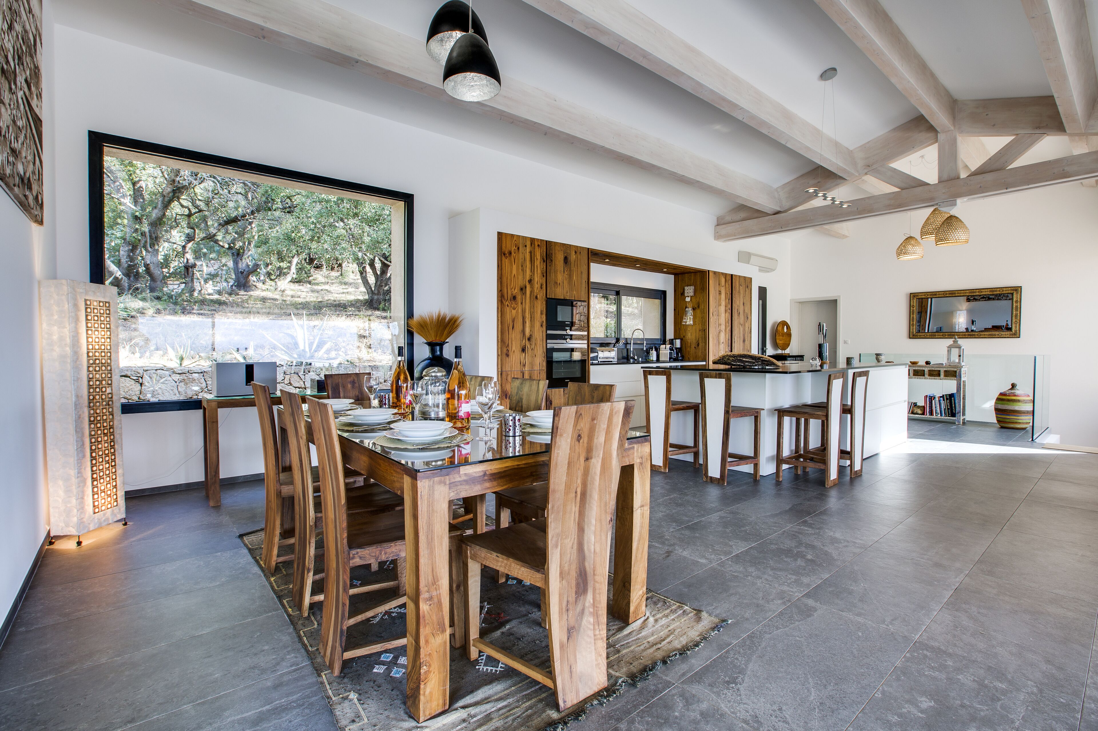 A spacious dining area with a large wooden table and chairs, a kitchen in the background, and a view of the outdoors through large windows.