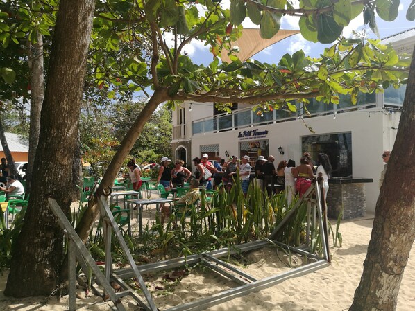 An outdoor seating area with tables and chairs, a building with a sign, and trees.