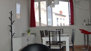 A dining area with a white table and chairs, a black chair in the foreground, and a view of a building through large windows with red curtains.