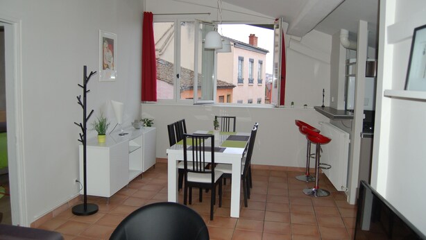 A compact kitchen with a dining area, a window with red curtains, and a view of buildings outside.
