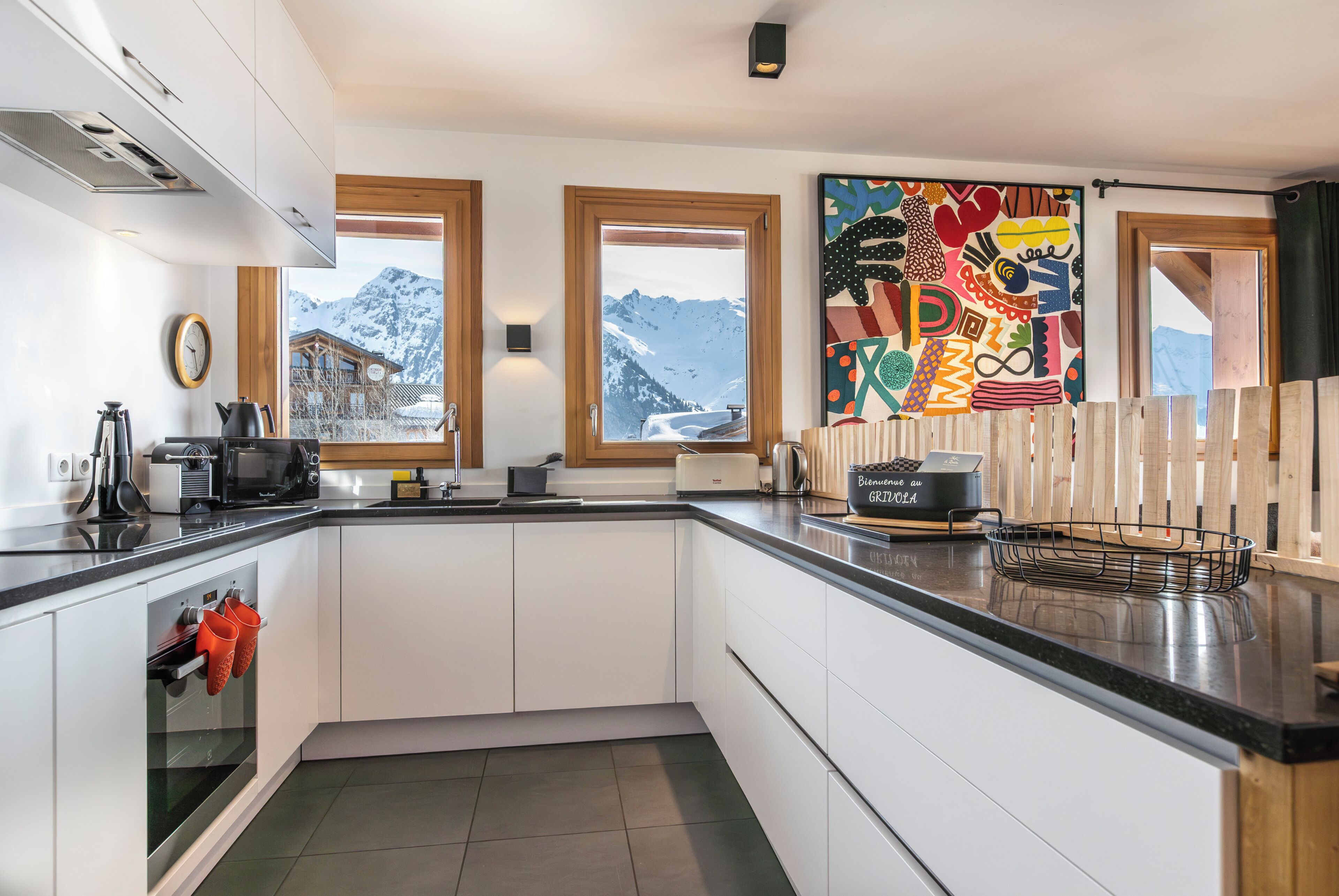 A modern kitchen with white cabinets, a black countertop, a wooden-framed window, and a colorful abstract painting.