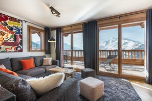 A modern living room with a sofa, ottoman, and a table, large windows with a mountain view, and a wooden deck outside. - 110m² apartment, 4 * full services, 4 bedrooms, 3 bathrooms / d, south balcony and 2 parking spaces (Montvalezan)
