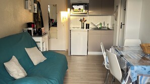 Un espai de viure compacte amb un sofà color turquesa, una taula de menjador amb cadires, una cuineta i un refrigerador petit.