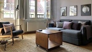 A modern living room with a grey sofa, a wooden coffee table, and a floor lamp. There are large windows with curtains, a radiator, and artwork on the wall. - Sunny Old Town Flat •  Walk Everywhere • Sleeps 4 (Bern)