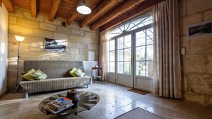 A cozy living room with a stone wall, wooden ceiling beams, a sofa with cushions, a glass coffee table with magazines, and a large window with curtains. - La Treille du Mas Vacation Rental—3-Star Rating (Arles)