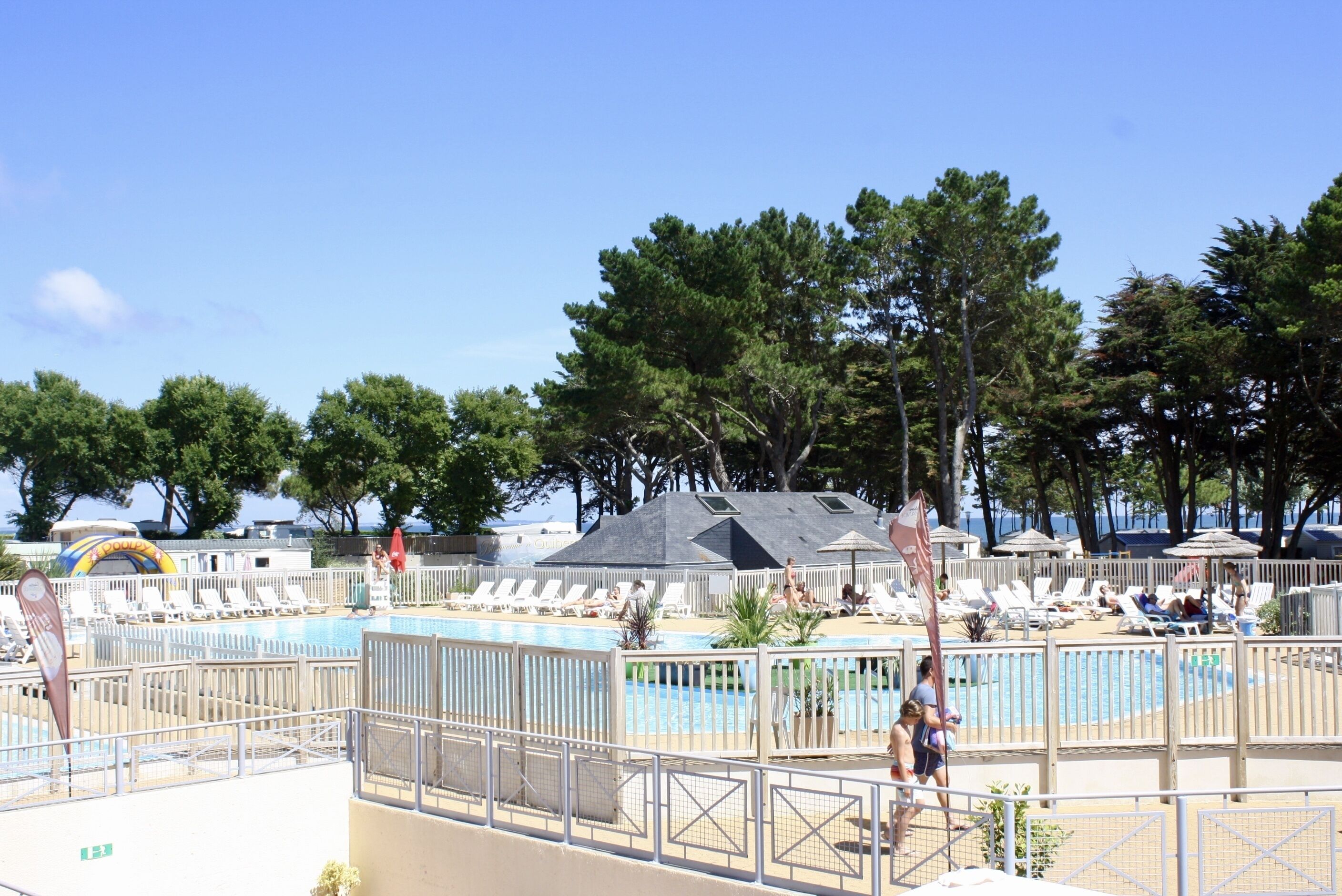 A pool area with lounge chairs and umbrellas, surrounded by trees and a building with a sloped roof.