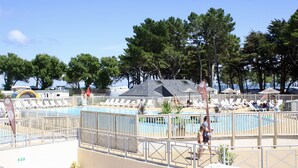 A pool area with lounge chairs and umbrellas, surrounded by trees and a building with a sloped roof. - "La Mer à L'Horizon" - Mobilhome 4P Conguel 4* - France (Quiberon)
