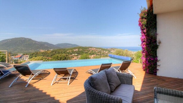 Una terraza de madera con sillones, piscina y vistas al mar y las colinas.
