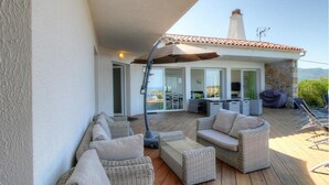 Un área de estar al aire libre techada con mobiliario de mimbre, una sombrilla y vista al interior a través de puertas de cristal.