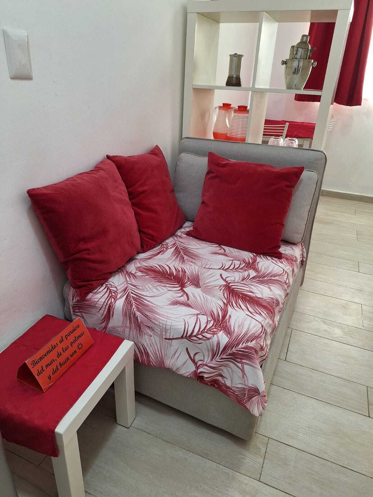 Ein kleiner, gemütlicher Raum mit einer grauen Schlafcouch, roten Kissen und einer floralen Tagesdecke. Vor der Couch steht ein kleiner Tisch mit einer roten Tischdecke und einem Willkommensschild.
