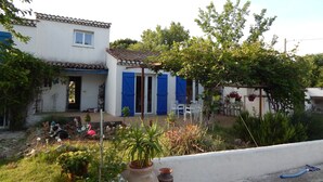 A white house with blue shutters, a garden with various plants and flowers, and a small patio area.