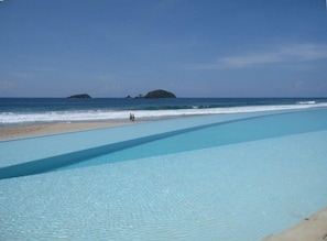 A clear blue pool adjacent to a sandy beach with a view of the ocean and distant islands.