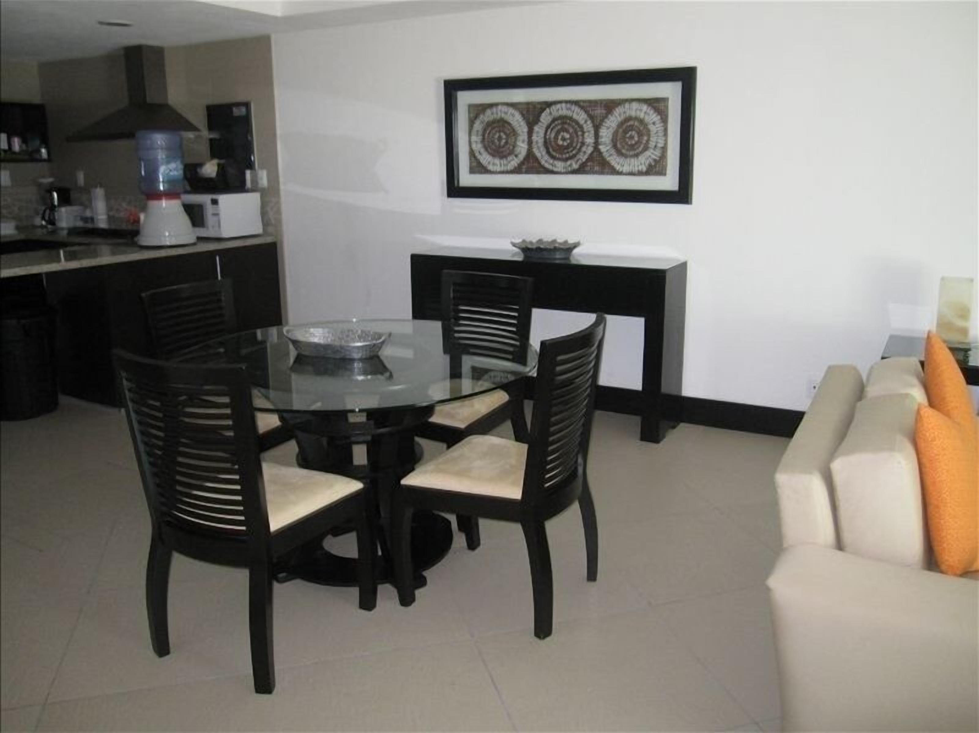 A dining area with a round glass table and black chairs, a kitchen in the background, and a framed artwork on the wall.