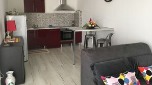 Uma cozinha compacta com forno embutido, uma área de jantar com mesa pequena e duas cadeiras, um sofá e um relógio fixado na parede.