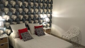 Um quarto com cabeceira estampada em padrão geométrico, cama com roupa de cama estampada, duas mesas de cabeceira com luminárias e uma cadeira branca.