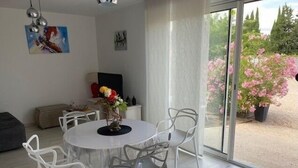 Uma área de refeições com uma mesa redonda e cadeiras brancas, um vaso de flores e vista para um jardim com flores cor-de-rosa através de uma porta de vidro.