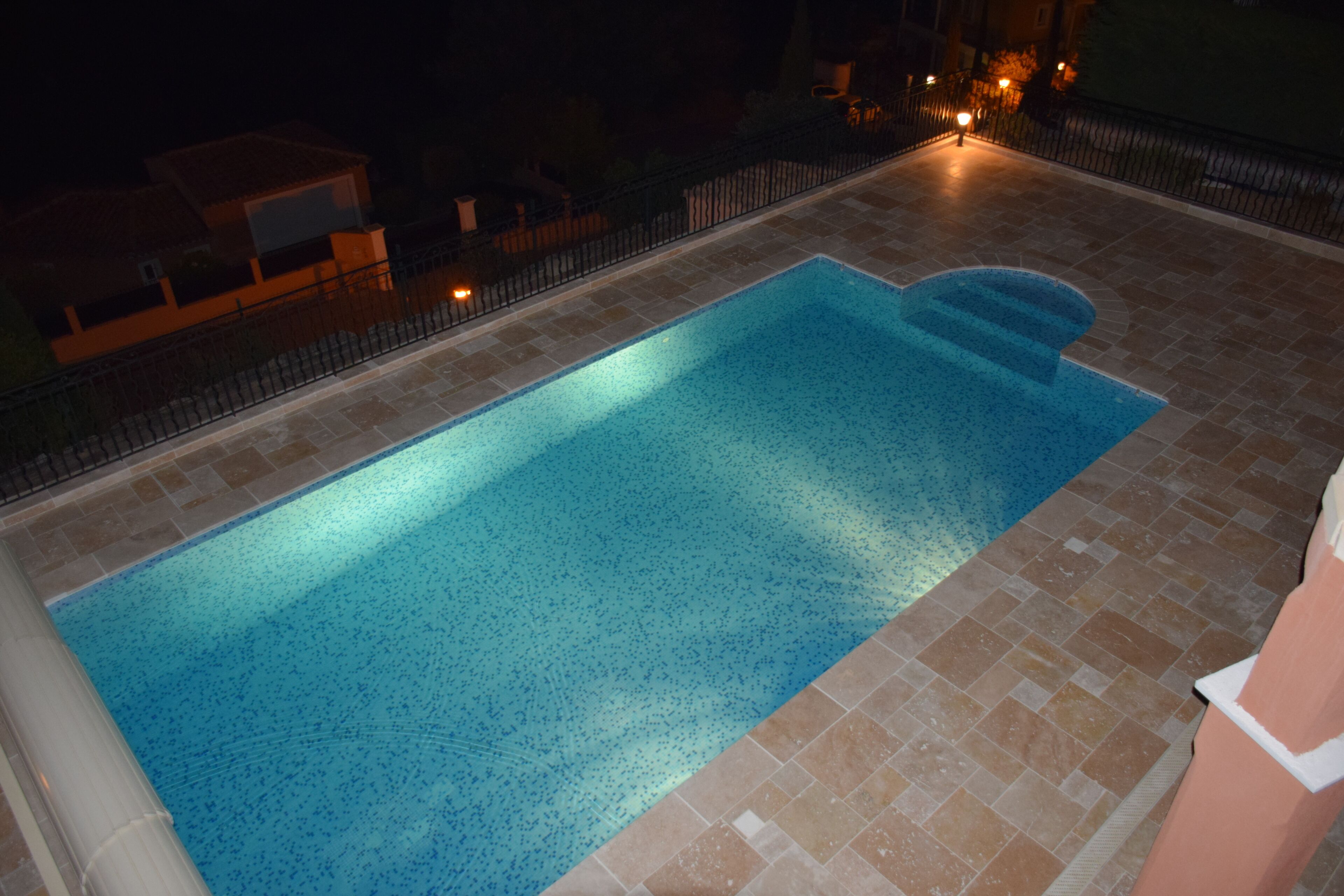 Ein rechteckiger Swimmingpool mit klarer, blauer Wasseroberfläche, umgeben von einer gefliesten Terrasse und einem schwarzen Metallzaun.