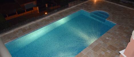 Ein rechteckiger Swimmingpool mit klarer, blauer Wasseroberfläche, umgeben von einer gefliesten Terrasse und einem schwarzen Metallzaun.