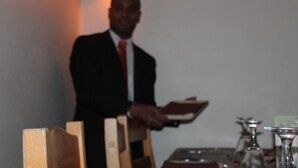 A person in a suit holding a tray, standing in a room with a table set for dining.
