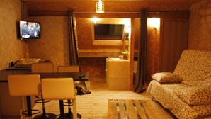 A cozy room with a wooden floor, a sofa, a dining table with chairs, a television, and a window with curtains.