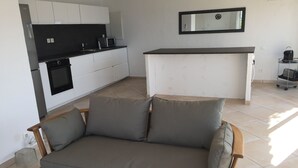 A modern kitchen with white cabinets, a black countertop, and a microwave. There is a grey sofa with wooden legs in the living area.
