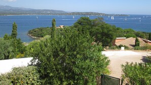 A coastal view with sailboats on the sea, greenery, and buildings in the background.