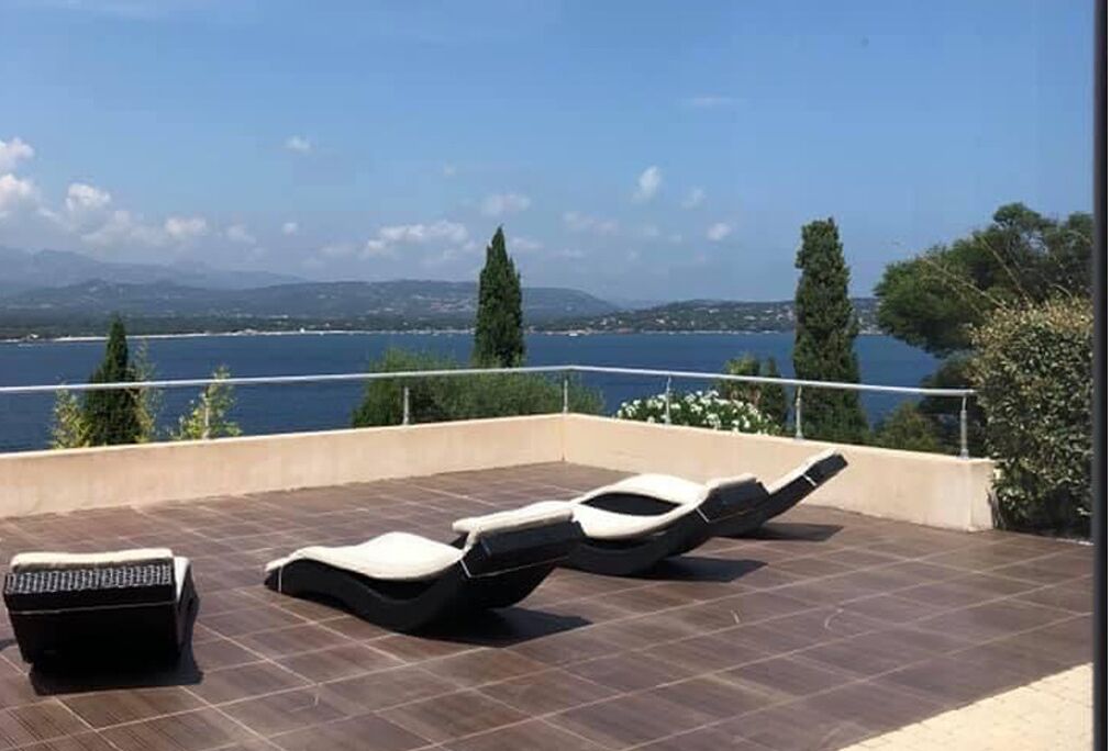 A rooftop terrace with three sun loungers and a small table, overlooking a body of water and distant hills.
