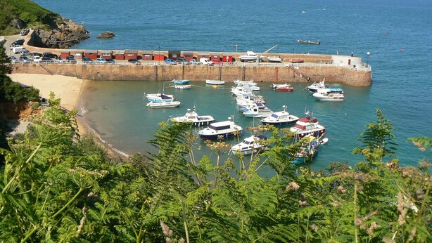 Una marina costera con barcos amarrados, un muro de piedra y una playa.