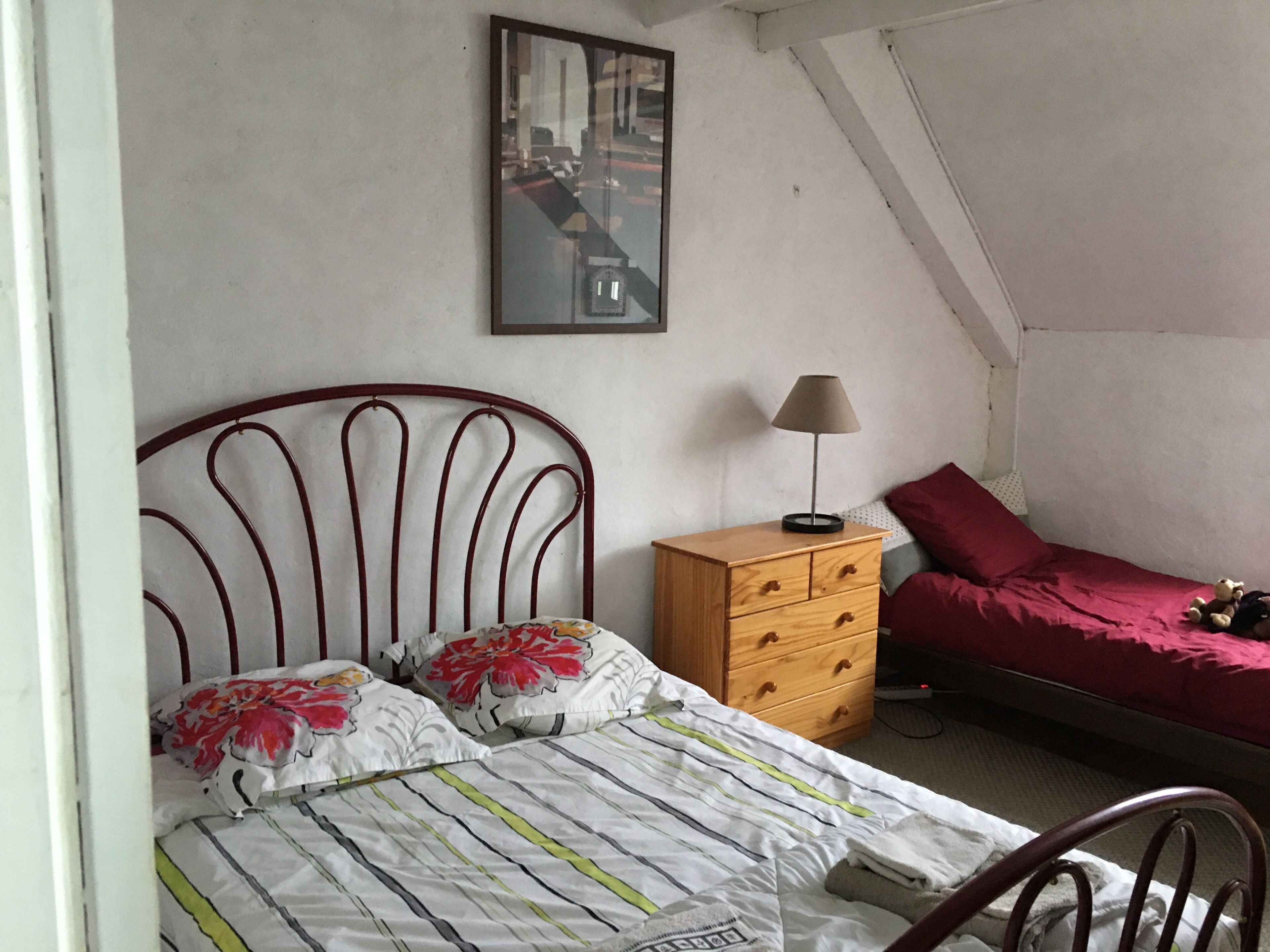 Un dormitorio con cama, cómoda, mesita de noche con lámpara, colcha roja y un cuadro en la pared.