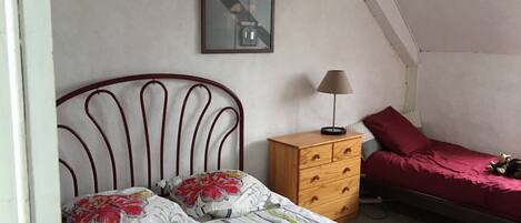 Un dormitorio con cama, cómoda, mesita de noche con lámpara, colcha roja y un cuadro en la pared.