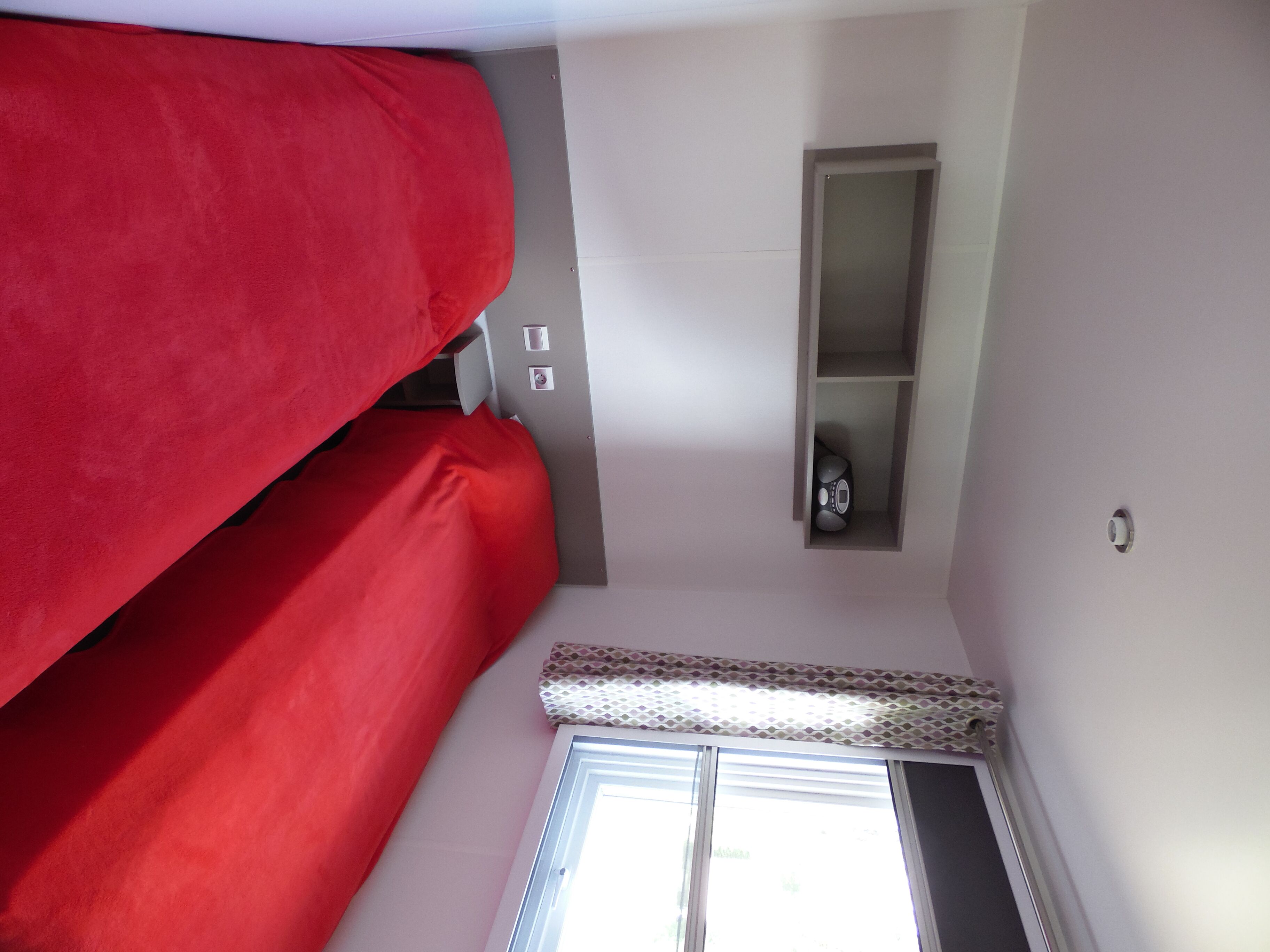 Ein kleiner Raum mit Etagenbett, einem kleinen Regal mit Lautsprecher und einem Fenster mit Jalousien.