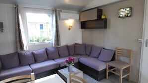A modern living room with a sectional sofa, a wooden chair, a small table, and a wall-mounted shelf. - BEAUTIFUL AIR-CONDITIONED MOBILE HOME RENTAL 6-8 PEOPLE LA PALMYRE AUX CHARMETTES 4E (Les Mathes)