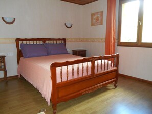 Una cama de madera con cabecera, dos almohadas, una mesita de noche, un reloj de pared y una ventana con cortinas.