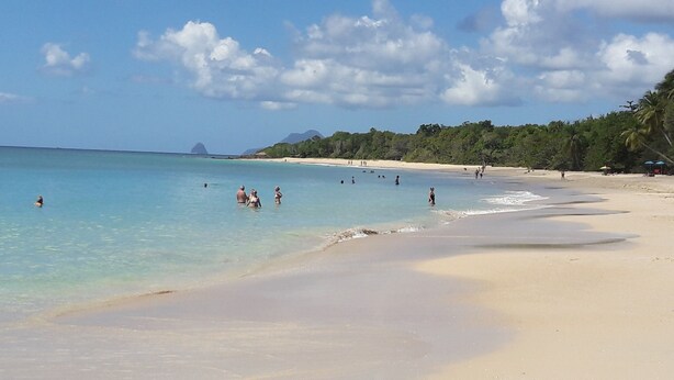 A beach with clear turquoise water, people swimming and walking along the shore, and a backdrop of green hills and a mountain.