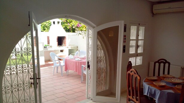 An open doorway leading to a patio area with a table and chairs, a brick oven, and a white wall with a window. - Ideal for holidays (NABEUL)