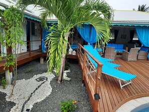 Eine Holzterrasse mit Liegestuhl und kleinem Tisch, umgeben von einem blauen Baldachin und einer Palme.