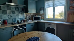A kitchen with a round wooden table, a microwave, and a window with blinds. - Homestay (Pontvallain)